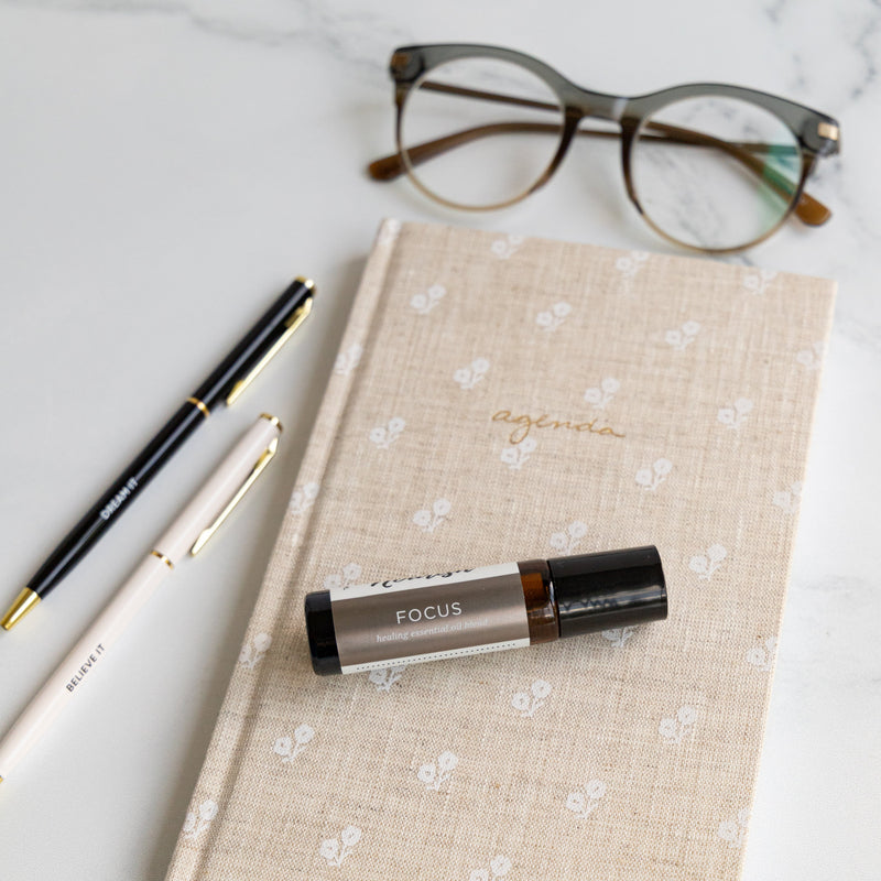 Notebook with a Focus Roller Blend, two pens, and glasses on a light surface.