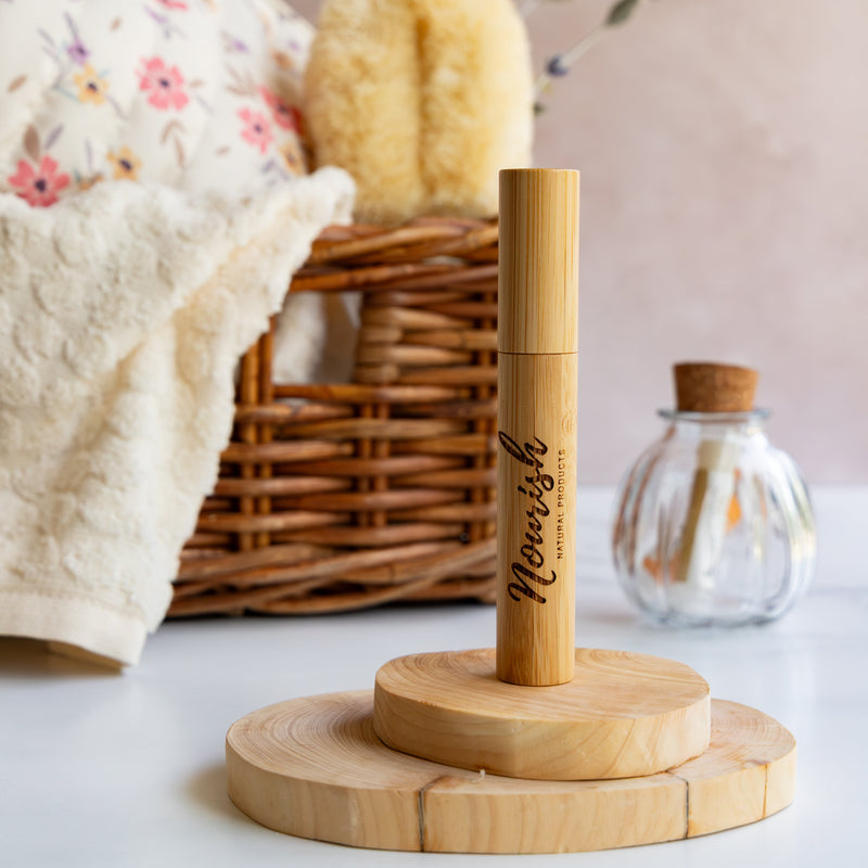 Wooden product stand with a Lash & Brow Growth Serum tube against a light surface with a basket and towel in the background