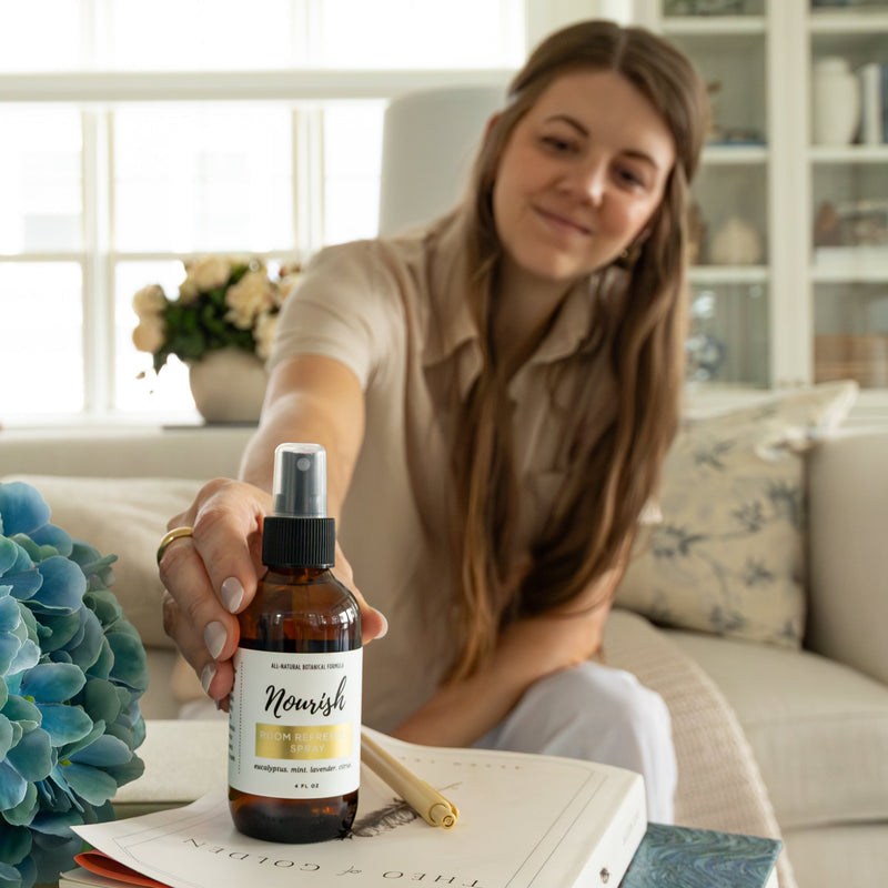 Woman holding a bottle of Room Refresher Spray in a home setting with a book and flowers on a table.