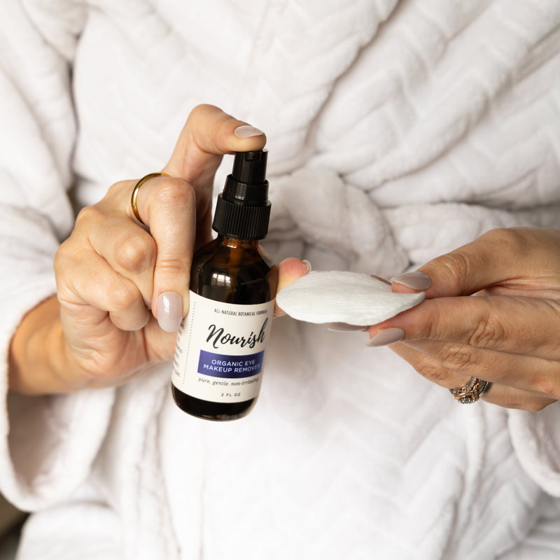 Person holding a bottle of Eye Makeup Remover and a cotton pad in a white robe.